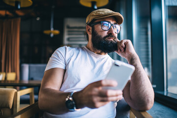 Fototapeta na wymiar Pensive caucasian bearded male using mobile phone spending free time in coffee shop dreaming about plans and ideas, handsome mature man in eyewear pondering and resting with smartphone in cafeteria