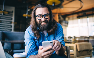 Happy hipster man in glasses using smartphone in modern cafe