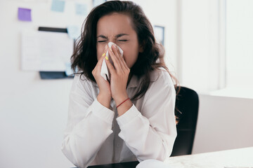 Sick and overworked businesswoman sitting at the desk in the office and blowing her nose, feeling unwell. Concept of hard work without maintaining health. Office work or work from home.