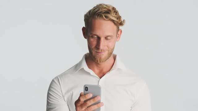 Handsome blond bearded man in shirt confidently using smartphone over white background. Young businessman working on phone