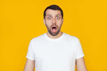 Obraz premium Young man with beard in white t-shirt with open mouth, afraid and shocked by unexpected expression, fear and excited face, stands on isolated yellow background