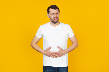A young man with a beard in a white T-shirt with a hand on his stomach because of indigestion, feeling sick. Pain concept. Stands on isolated yellow background