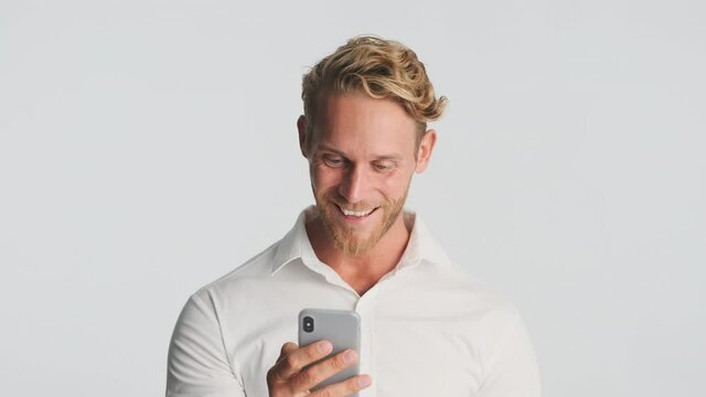 Handsome smiling blond bearded businessman in shirt happily using smartphone over white background