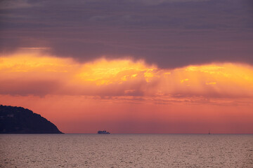 Mediterranean sea sunset during a storm. Orange sun rays filtered by the dark clouds. Tip of an island on the left side. A cruise boat navigates at the horizon.