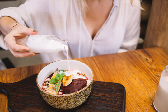Cropped image of female holding bottle with milk pouring bowl with granola having healthy meal in morning, woman enjoying oat cereals with fruits and cream having sweet brunch appetizer