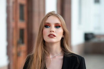 A girl with red hair, dressed in a black jacket, posing against the backdrop of a white building. Looking away.