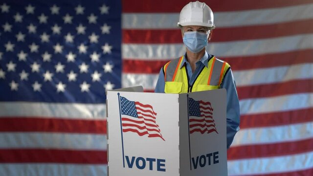 Conceptual portrait US flag background with smiling woman engineer architect construction worker face mask giving thumbs up in voting booth at ballot box, careful decision for president US election.