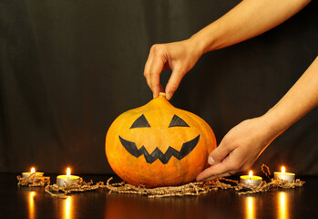 Mother's hands put a pumpkin with a face made by child on the table. The child's hand gives a burning candle. Process of decoration of home for Halloween party. Copy space