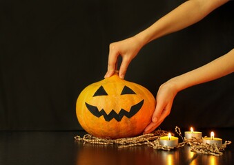 Child's hands put a pumpkin with handmade face on the black table next to burning candles. The process of decoration of home for Halloween party.  Copy space