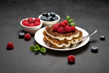 portion of Classic tiramisu dessert with raspberries and blueberries on dark concrete background
