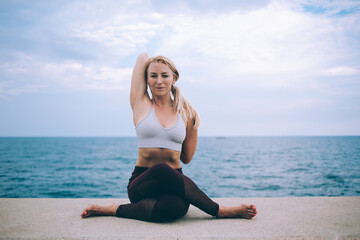 Fototapeta na wymiar Portrait of Caucasian female yogi sitting in Gomukhasana pose and looking at camera during morning workout near sea,experienced woman in tracksuit practicing physical exercise during energetic pilates