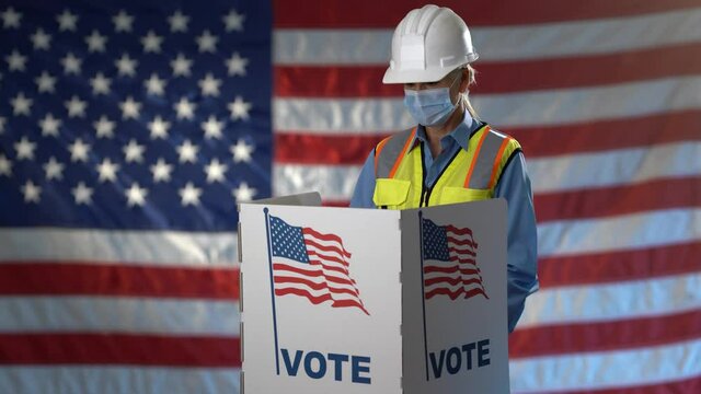 Conceptual US flag background with smiling woman engineer architect construction worker face mask in voting booth at ballot box, careful decision in US election.