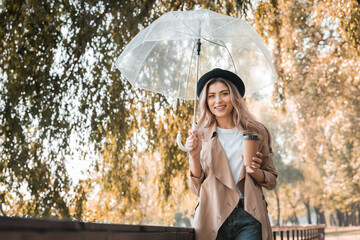 blonde woman in hat holding umbrella and paper cup with coffee to go in autumnal park