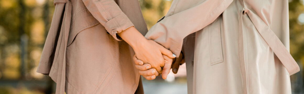 panoramic crop of man and woman in trench coats holding hands in park