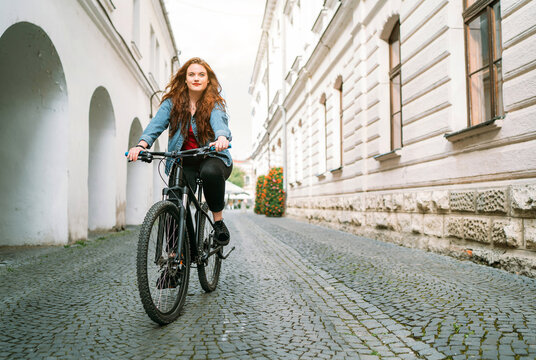 The portrait of Red curled long hair caucasian teen girl on the cobblestone city street riding a bicycle. Natural people beauty urban life concept image.