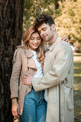 pleased man with closed eyes hugging girlfriend in trench coat in autumnal park