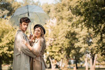 blonde woman in hat and man in trench coat hugging under umbrella and looking at camera in autumnal park