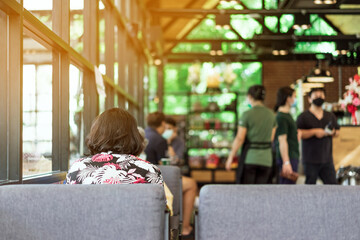 Naklejka premium Back view of Asian female customer wear face mask using smartphone while sit in waiting social distance queue for take away drink in coffee shop. New normal lifestyle concept.