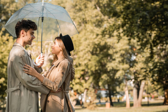 Side View Of Woman In Hat And Man In Trench Coat Standing Under Umbrella In Autumnal Park