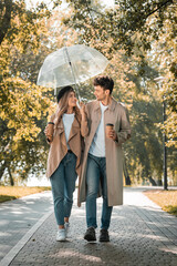 couple in trench coats walking under umbrella and holding paper cups with coffee to go