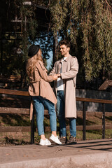 Fototapeta na wymiar joyful man in trench coat looking at woman in hat while holding hands in park