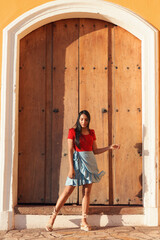Young female on a summer day wearing a red shirt, bright blue skirt and beige espadrilles sandals posing next to a big vintage door