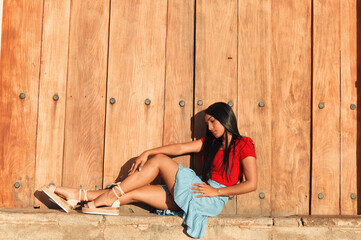 A young female on a summer day wearing a red shirt, bright blue skirt and beige espadrilles sandals posing sitting next to a big vintage door