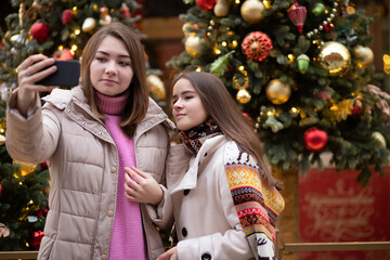 Christmas mood. Young girls take a selfie on a smartphone against the background of lights, Christmas trees, toys, New Year's fair.