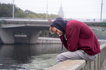 Portrait of a young unshaven sad pensive guy sitting on a fence near the river. Teenager looking at the river