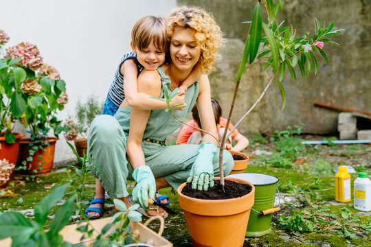 Cute Child Boy Hugging His Mother Gardener At Backyard Garden. Happy Lovely Mom With Children Outdoor. Hobbies And Leisure, Home Gardening, Houseplant, Urban Jungle Concept.