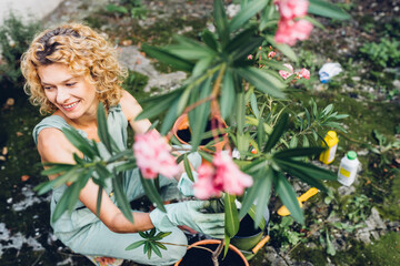 View from above portrait of happy middle age blond woman with blooming oleander plant in pot in backyard or garden. Female gardener planting plants in pots.