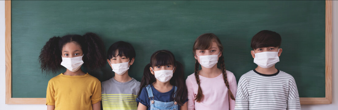 Portrait Of Diversity Elementary School Pupils with protective face mask on blackboard in classroom.