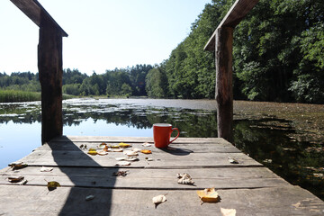 Orange mug with tea among the autumn leaves on the bridge on the background of a forest lake, side view-the concept of a pleasant holiday in autumn