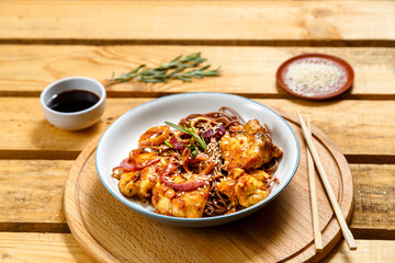 plate of soba with chicken and sesame seeds in teriyaki sauce on a wooden stand and sticks next to sesame seeds and soy sauce.