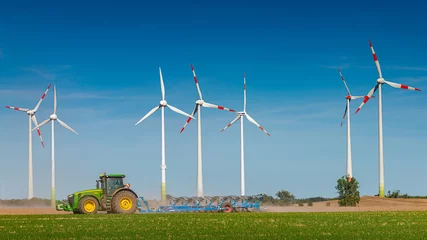 Fototapeten Große Windkraftanlagen zur Stromerzeugung, grüne, umweltfreundliche Energie am blauen Himmel, die auf dem grünen Feld des Bauernhofs stehen, mit einer funktionierenden Landmaschine als Traktor darin, bei warmen Sonnenuntergangsfarben ... © neurobite