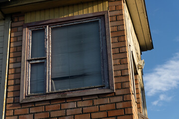 Old brick house with a yellow roof and an old gray wooden window against the blue sky.