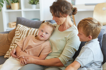 Obraz premium Portrait of modern mother embracing girl with down syndrome while sitting on couch with two kids in home interior