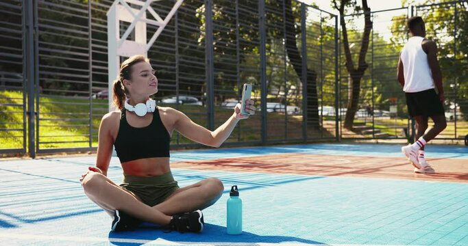 Beautiful Caucasian cheerful female athlete sitting on basketball court and making selfie photo on smartphone on sunny day. African American male playing streetball on background. Leisure concept