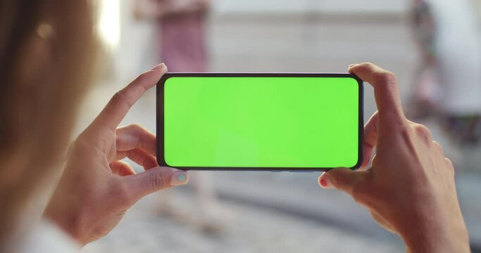 Close up view of female hand holding smartphone with mockup template screen horizontally. Concept of greenscreen and chroma key. People passing by at background.