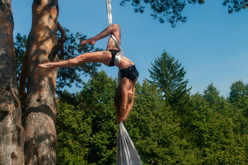A slender girl with blonde hair, in a black stage costume, performs gymnastic and circus exercises on white silk against the background of the forest and the sky. © lik13vvs