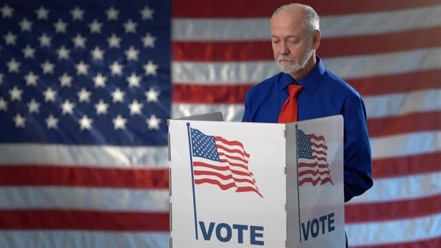 Conceptual portrait on US flag background of smiling man in voting booth at ballot box, careful decision for president, with thumbs up for US election during pandemic.