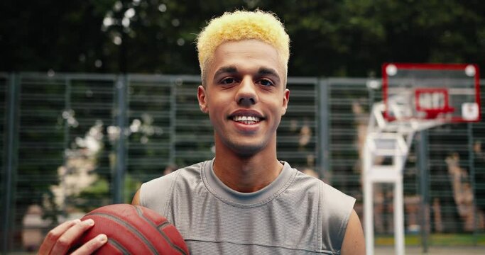 Close up portrait of young happy guy standing on urban basketball court with ball outdoors. Joyful male streetball player smiling to camera on street. Sporty lifestyle. City concept