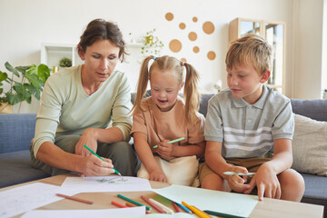 Portrait of cute blonde girl with down syndrome laughing happily while drawing together with mother and brother at home