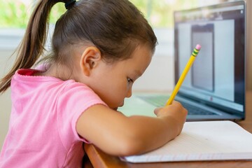 A kindergarten student learning how to write with pencil and paper, online school. Distance learning.