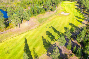 Aerial view down on unidentified people play golf on Golf course, surrounded by pine tree forest in Scandinavia. Warm Sunny calm day excellent for golfers. Narrow long golf course in Northern forest
