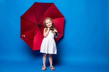 Happy little girl with red umbrella posing on blue wall background.