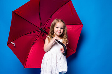 Happy little girl with red umbrella posing on blue wall background.