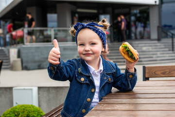 Little cute girl eating a burger showing thumbs up in a cafe. Concept of a children's fast food meal