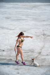 A red-haired woman in a bikini walks with a dog on a snowy beach