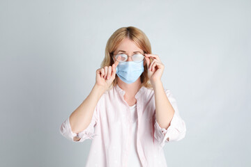 Woman wiping foggy glasses caused by wearing medical mask on light background